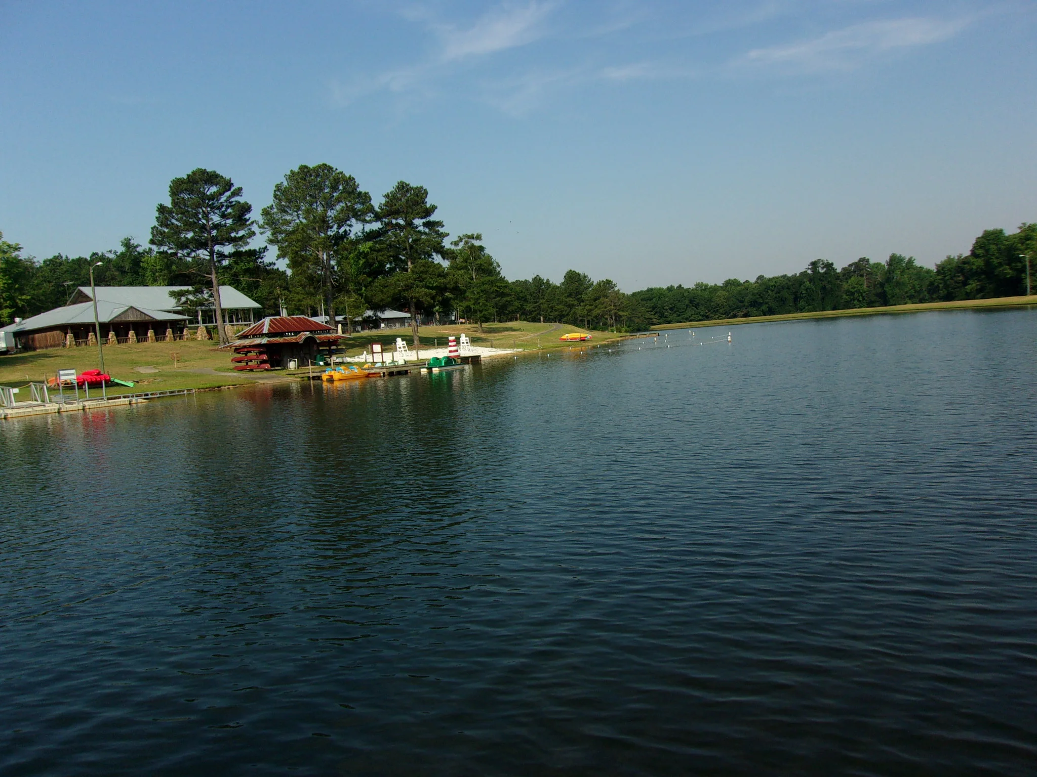 Calm lake shoreline with a wooden boathouse and docked kayaks, surrounded by leafy trees under a soft blue sky, peaceful and welcoming.