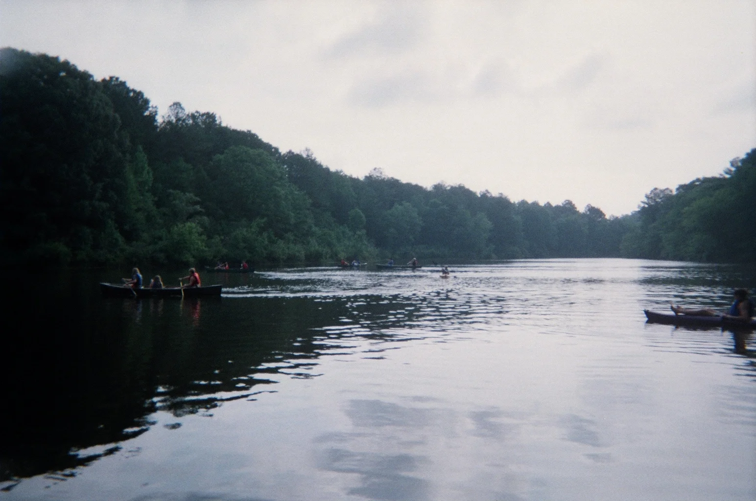 A calm lake flanked by tree-covered hills with canoes gliding on the surface, warm late afternoon light and muted greens and earth tones.