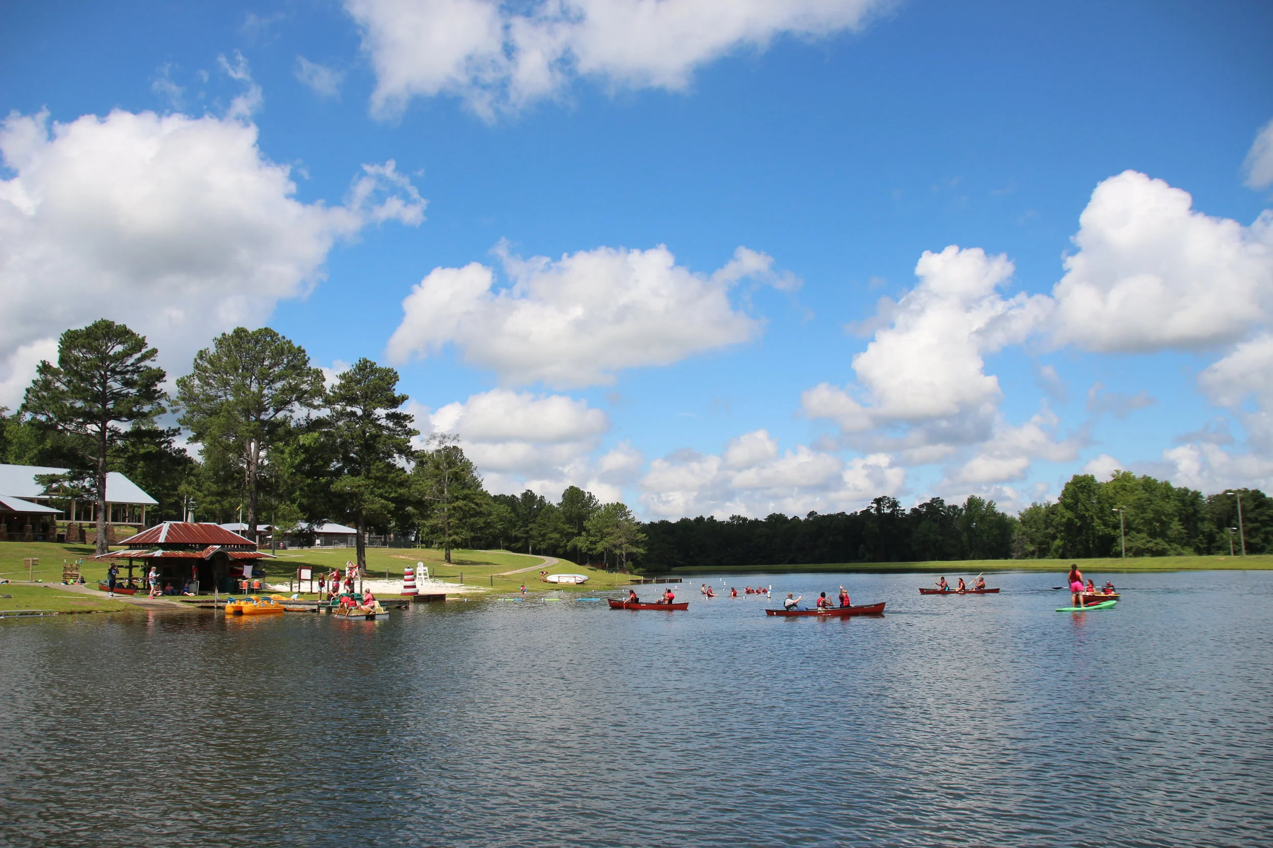 Wide outdoor view of a lake with canoes and kayaks near a dock and a lakeside pavilion, blue sky with soft clouds.